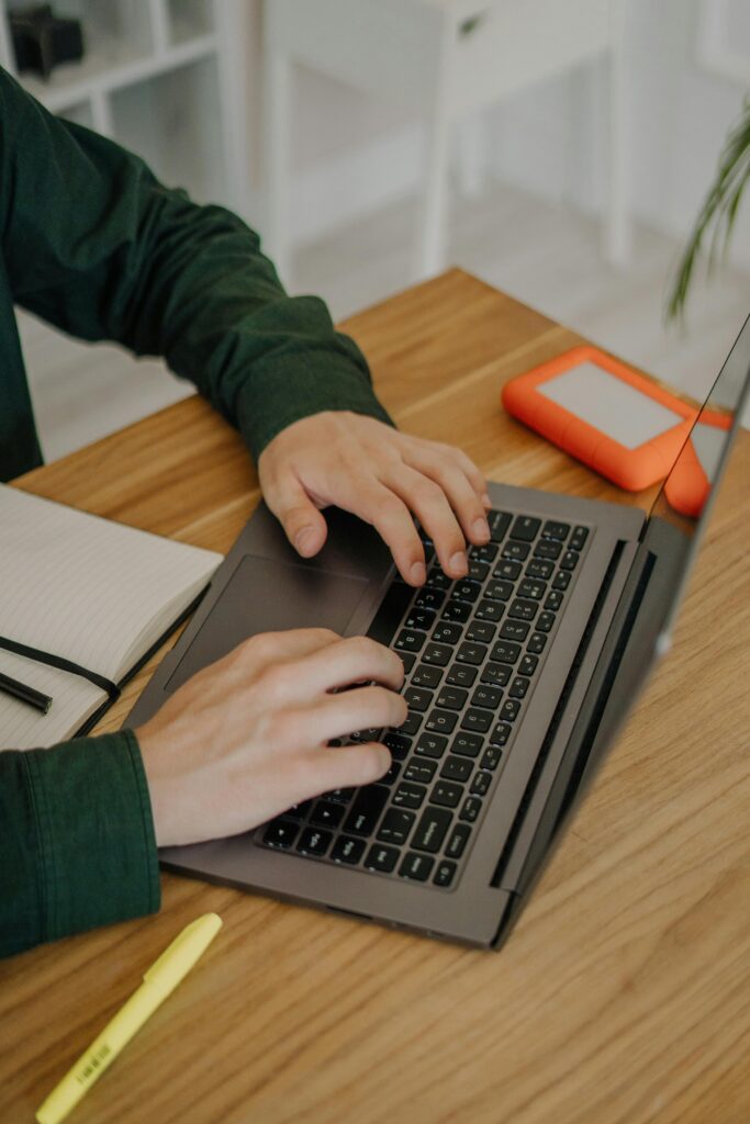 Hands typing on a laptop at a wooden desk with an external hard drive nearby.